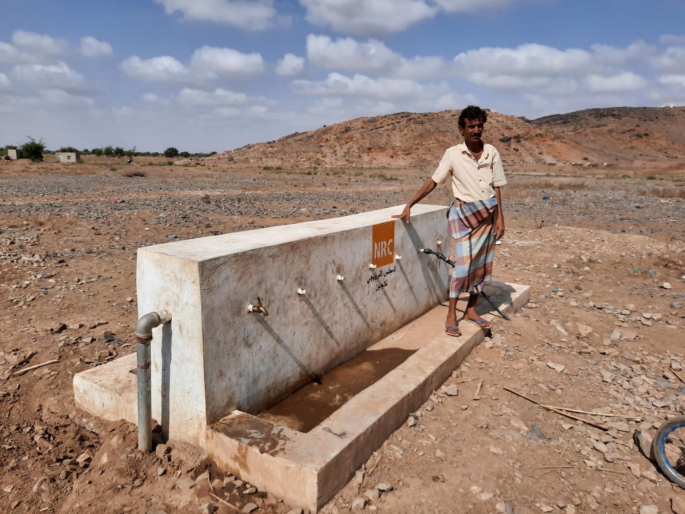 Hassan Abdullah Ahmed Abdu, aged 36, is originally from Hajjah’s Haradh district. He fled his home—together with his family and all their neighbours—when the conflict reached their village in 2015. Like many other displaced people, he left in a hurry and only managed to take the most important things with him, like documents.

Hassan is a father of five children, and he used to work at many jobs, including selling Qat. But after he left his village, he could not find another job. He is now dependent on humanitarian assistance to provide for his family.

Hassan arrived in Shib Al-Argah village, in Al-Hodeidah’s Az-Zuhrah district. where many other displaced families live inside Oshas (straw huts) and since 2015 he has been suffering to provide his family with basic needs.

“Since we arrived this area, I have been suffering. Providing my family with food and other basic services and obtaining water was the main challenge for us,” he says. “My family and other families in this village used to walk for two kilometres to fetch water from the well, [carrying it] either on their heads or on donkeys. That was real suffering.”

“My family and other families in this village used to walk for two kilometres to fetch water from the well, [carrying it] either on their heads or on donkeys. That was real suffering.”

Thanks to the support of UK Aid, the Norwegian Refugee Council (NRC) intervened and ended this suffering. First, NRC constructed masonry tanks and installed pumps. Distribution pipes were then added, and 11 water distribution points constructed.

When NRC began the project, Hassan helped directly with the construction. As part of the cash-for-work team, he worked on the excavation and backfilling works to extend the water pipes to provide clean water.

“After the completion of the water project, conditions have become better,” Hassan says. The new pipes bring clean water directly to key water points where families are gathered. “Our difficulty getting clean water was completely solved,” he adds.

Hassan is completely satisfied with the project and also feels great happiness now that it has finished, as it has alleviated a burden from the community. This is especially true for women and children, who usually had the task of collecting the water. Now they can gather the water they need daily without any trouble.

“It is good that we have enough water and we have started to adapt to the life in the camp,” Hassan concludes, “but my dream is still to return my village. I hope the war ends and I can return.”

Photo: Ahmed Ibrahim/NRC