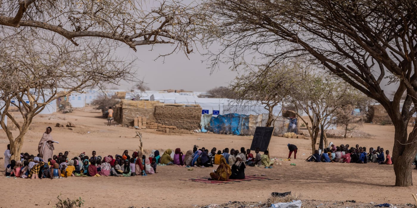 En grupp kvinnor och barn sitter samlade i skuggan av några träd i ett torrt och dammigt landskap. I bakgrunden syns enkla skydd byggda av lera och presenningar i ett flyktingläger.
