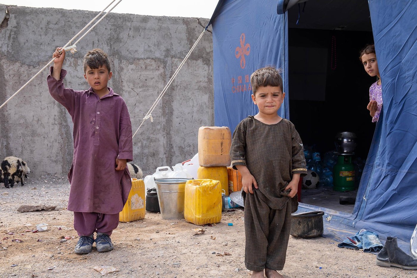 Three siblings outside the tent they are living in. The boys are outside, while their sister is looking out from inside the tent.