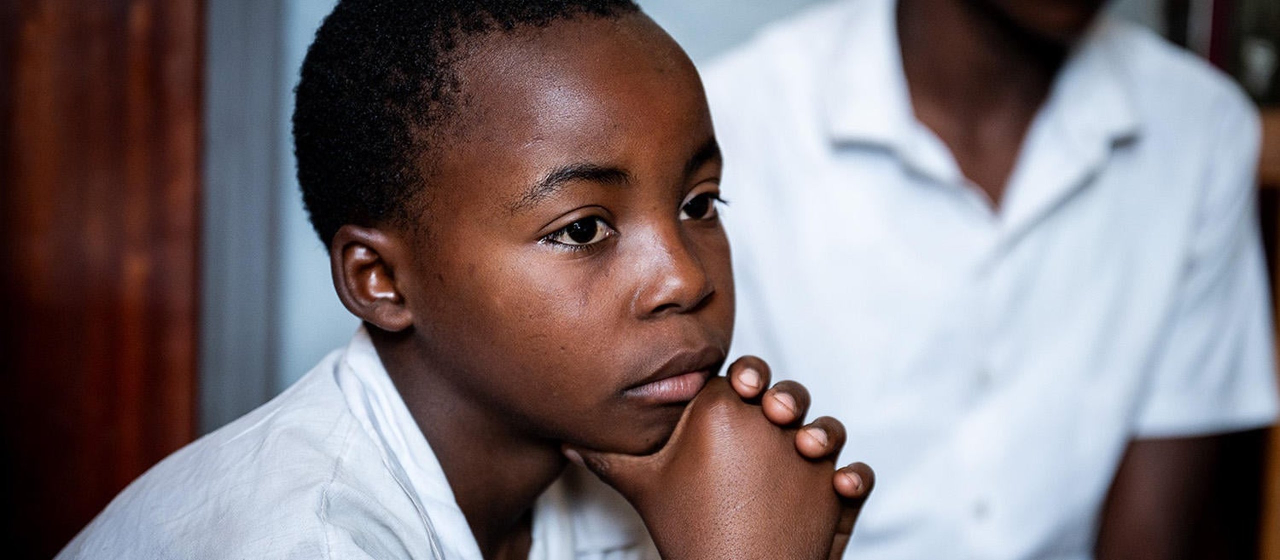 A boy resting his chin in his hands, looking at something to the right of the photos edge. He is wearing a white school uniform shirt.