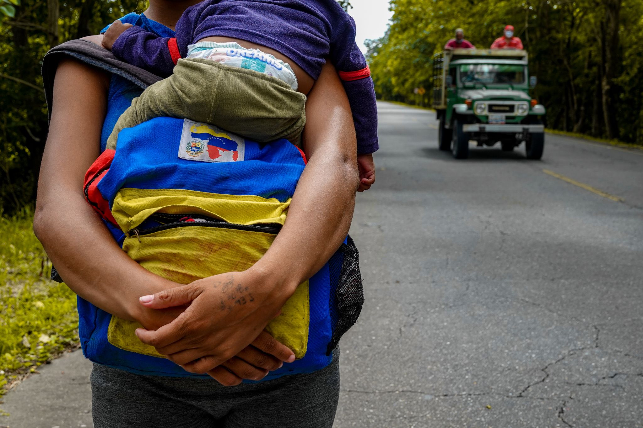 A person on the road, holding a child and a backpack. Behind them is an open-top car with two people.